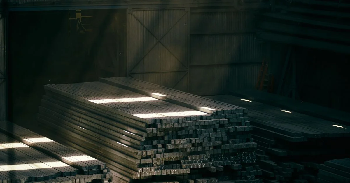 Des piles de poutres métalliques dans l'intérieur d'un entrepôt industriel baigné de soleil.