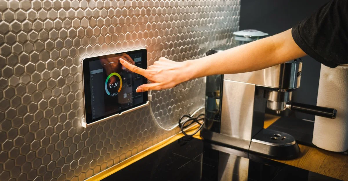 Scène de cuisine moderne avec une main utilisant une tablette pour contrôler un système de maison intelligente.