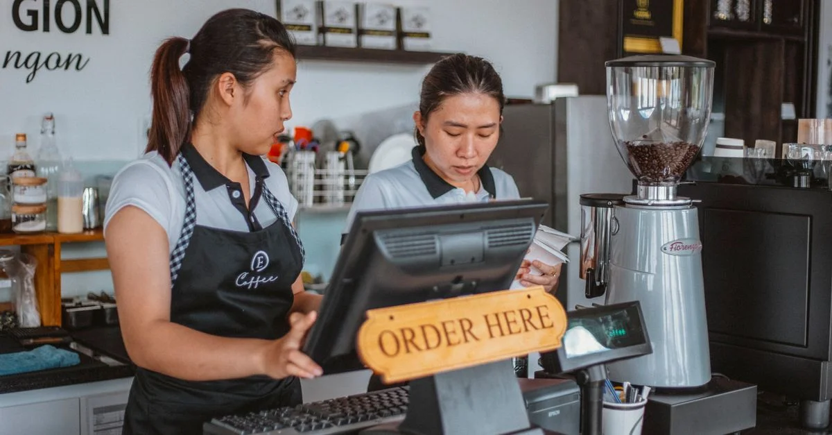 Dos baristas trabajan en el mostrador de una cafetería con una caja registradora y equipos.